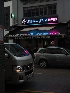 A vibrant bistro pub with a neon sign displaying colorful text in both English and another language. The establishment offers local and western food and advertises live football. Parked in front of the pub are two vehicles, a white van and a silver car, on a dimly lit street.
