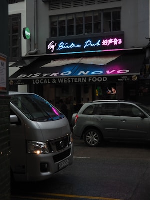 A vibrant bistro pub with a neon sign displaying colorful text in both English and another language. The establishment offers local and western food and advertises live football. Parked in front of the pub are two vehicles, a white van and a silver car, on a dimly lit street.