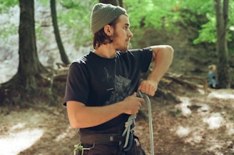 A person wearing a beanie and a dark t-shirt stands in a forested area, holding a coiled rope in their hands. The surrounding environment is lush with green foliage and trees with visible roots. The background is slightly blurred, showing another person in the distance who is also in the forest.