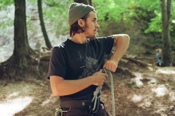 A person wearing a beanie and a dark t-shirt stands in a forested area, holding a coiled rope in their hands. The surrounding environment is lush with green foliage and trees with visible roots. The background is slightly blurred, showing another person in the distance who is also in the forest.