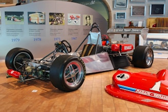 A partially assembled race car with visible chassis and engine parts is displayed in an exhibition setting. The rear tires are large and fitted for racing, and a red Renault logo is noticeable on the side. Nearby, a bright red car front with the number '1' is positioned on the wooden floor. The wall behind features a timeline with images and text relating to vehicle design and construction over the years.