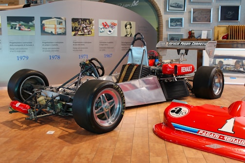 A partially assembled race car with visible chassis and engine parts is displayed in an exhibition setting. The rear tires are large and fitted for racing, and a red Renault logo is noticeable on the side. Nearby, a bright red car front with the number '1' is positioned on the wooden floor. The wall behind features a timeline with images and text relating to vehicle design and construction over the years.