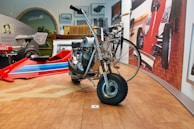 A vintage motorcycle with a sidecar is displayed prominently. Surrounding it are various car models and automobile-related paraphernalia. The room features a polished wooden floor and several framed photographs and artworks on the walls.