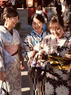 Three women wearing colorful kimonos are walking outside. They are smiling and appear to be enjoying each other's company. The kimonos have various floral and geometric patterns. They are carrying small bags, and the setting seems to be sunny with people and steps in the background.