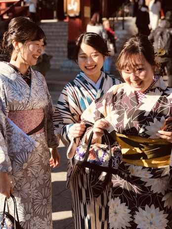 Three women wearing colorful kimonos are walking outside. They are smiling and appear to be enjoying each other's company. The kimonos have various floral and geometric patterns. They are carrying small bags, and the setting seems to be sunny with people and steps in the background.