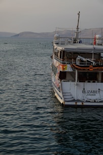 A large boat named 'Elizabeth Zadar' is docked in calm waters. The boat's hull features colorful designs, and it has multiple decks including visible safety equipment. The backdrop includes a serene expanse of water extending towards a mountainous horizon under a light sky.