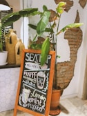 A cozy cafe setting features a wooden-framed chalkboard displaying a handwritten menu with items like frappe, strawberry matcha, lemon mint, and mojito. Surrounding the chalkboard are potted plants with broad green leaves, enhancing the warm and inviting atmosphere. A rustic wall with exposed brick adds to the charm.