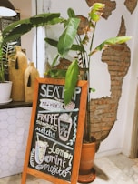 A cozy cafe setting features a wooden-framed chalkboard displaying a handwritten menu with items like frappe, strawberry matcha, lemon mint, and mojito. Surrounding the chalkboard are potted plants with broad green leaves, enhancing the warm and inviting atmosphere. A rustic wall with exposed brick adds to the charm.