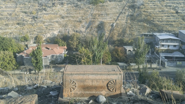 A scenic landscape shows a grassy hillside with terraced earth, small trees, and residential buildings with red and gray rooftops. In the foreground, there's a stone structure with engraved writing.