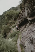 A person is walking on a narrow, rocky trail surrounded by lush greenery and steep rock formations. The path winds through a hilly landscape with dense foliage and trees. The atmosphere is serene, natural, and isolated.