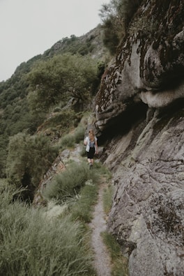 A person is walking on a narrow, rocky trail surrounded by lush greenery and steep rock formations. The path winds through a hilly landscape with dense foliage and trees. The atmosphere is serene, natural, and isolated.