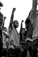 A group of individuals raise their fists in a gesture of solidarity and protest. The image is in black and white, emphasizing the serious and unified mood. The individuals appear focused and determined, each with an arm raised high.