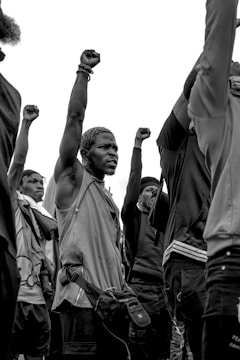 A group of individuals raise their fists in a gesture of solidarity and protest. The image is in black and white, emphasizing the serious and unified mood. The individuals appear focused and determined, each with an arm raised high.