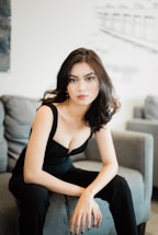 A woman with long, wavy hair is seated on a couch. She is dressed in a black outfit and gazes directly at the camera with a neutral expression. The background is blurred, showing parts of a room with modern decor.