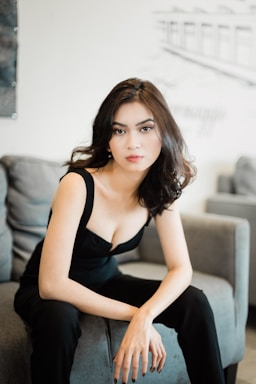 A woman with long, wavy hair is seated on a couch. She is dressed in a black outfit and gazes directly at the camera with a neutral expression. The background is blurred, showing parts of a room with modern decor.