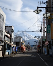A quiet street scene in Japan with overhead wires and signage lining the buildings. The street is empty, and in the background, a snow-capped mountain dominates the view.