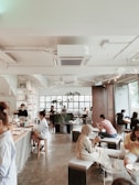 A modern cafe with a spacious, airy interior featuring tall windows allowing natural light to flood the space. Several people are sitting at various tables, engrossed in their activities, some using laptops or phones. The decor includes minimalist furniture and indoor plants, creating a relaxed and inviting atmosphere.
