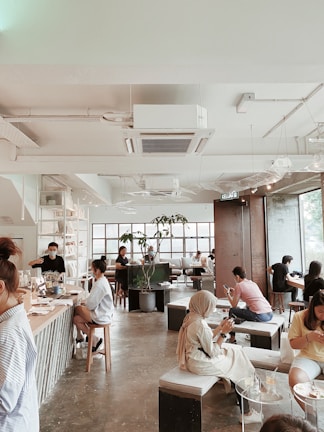 A modern cafe with a spacious, airy interior featuring tall windows allowing natural light to flood the space. Several people are sitting at various tables, engrossed in their activities, some using laptops or phones. The decor includes minimalist furniture and indoor plants, creating a relaxed and inviting atmosphere.