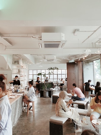 A modern cafe with a spacious, airy interior featuring tall windows allowing natural light to flood the space. Several people are sitting at various tables, engrossed in their activities, some using laptops or phones. The decor includes minimalist furniture and indoor plants, creating a relaxed and inviting atmosphere.