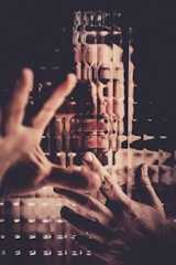 A person is seen through a textured, distorted surface with hands pressed against it. The image gives an impression of struggle or confinement, with the figure's facial expression suggesting distress or frustration.