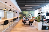 A modern coworking space with a stylish and open layout. The room features a long bar-style table with high black chairs, pendant lighting, and a glass partition on one side. Two people are seated at a round table in the center, engaged in discussion, and using laptops. The room is decorated with plants, shelves, and a large green abstract mural on the wall.