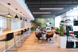 A modern coworking space with a stylish and open layout. The room features a long bar-style table with high black chairs, pendant lighting, and a glass partition on one side. Two people are seated at a round table in the center, engaged in discussion, and using laptops. The room is decorated with plants, shelves, and a large green abstract mural on the wall.