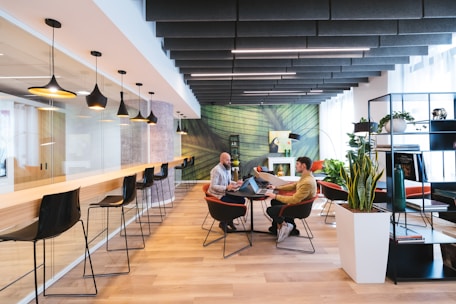 A modern coworking space with a stylish and open layout. The room features a long bar-style table with high black chairs, pendant lighting, and a glass partition on one side. Two people are seated at a round table in the center, engaged in discussion, and using laptops. The room is decorated with plants, shelves, and a large green abstract mural on the wall.
