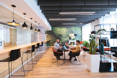 A modern coworking space with a stylish and open layout. The room features a long bar-style table with high black chairs, pendant lighting, and a glass partition on one side. Two people are seated at a round table in the center, engaged in discussion, and using laptops. The room is decorated with plants, shelves, and a large green abstract mural on the wall.