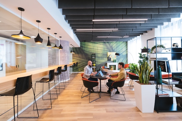 A modern coworking space with a stylish and open layout. The room features a long bar-style table with high black chairs, pendant lighting, and a glass partition on one side. Two people are seated at a round table in the center, engaged in discussion, and using laptops. The room is decorated with plants, shelves, and a large green abstract mural on the wall.