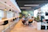 A modern coworking space with a stylish and open layout. The room features a long bar-style table with high black chairs, pendant lighting, and a glass partition on one side. Two people are seated at a round table in the center, engaged in discussion, and using laptops. The room is decorated with plants, shelves, and a large green abstract mural on the wall.