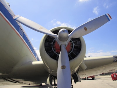 Close-up view of an airplane propeller and engine with a blue sky background. The propeller blades are metallic with a slight sheen, and the engine casing is cylindrical with visible bolts and components. Part of the aircraft's fuselage features a striped design.