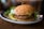 A close-up view of a veggie burger placed on a ceramic plate. The burger consists of a toasted bun with a patty that looks like it contains a mix of vegetables, topped with a green spread that resembles guacamole. A pickle slice and some greens are visible beside the burger on the plate. The background is softly blurred, emphasizing the burger in focus.