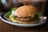 A close-up view of a veggie burger placed on a ceramic plate. The burger consists of a toasted bun with a patty that looks like it contains a mix of vegetables, topped with a green spread that resembles guacamole. A pickle slice and some greens are visible beside the burger on the plate. The background is softly blurred, emphasizing the burger in focus.