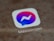 A close-up view of a smartphone screen displaying the Messenger app icon, which is a speech bubble with a gradient from blue to pink and a white lightning bolt in the center.
