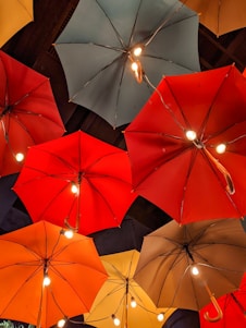 A collection of umbrellas is arranged in an overhead display, each fitted with small light bulbs, creating an artistic and whimsical atmosphere. The umbrellas are of various colors, including red, orange, yellow, and teal, and hang from the ceiling in an artistic manner.