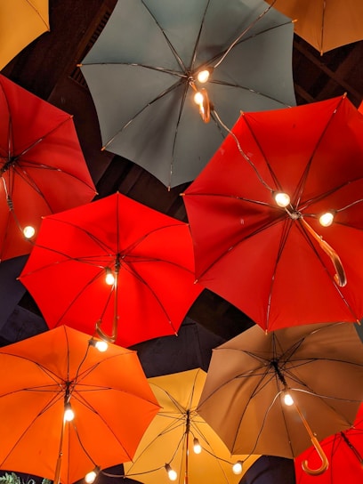 A collection of umbrellas is arranged in an overhead display, each fitted with small light bulbs, creating an artistic and whimsical atmosphere. The umbrellas are of various colors, including red, orange, yellow, and teal, and hang from the ceiling in an artistic manner.