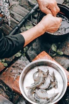 Hands preparing prawns over a pot, with a bowl containing prawns nearby, set on a rustic outdoor surface with bricks and scattered leaves.