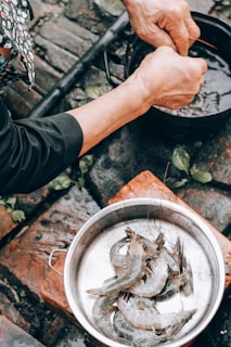 Hands preparing prawns over a pot, with a bowl containing prawns nearby, set on a rustic outdoor surface with bricks and scattered leaves.