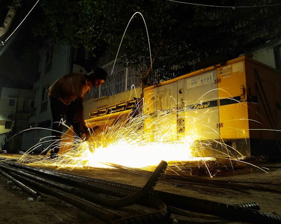 A person is using a welding or cutting tool, generating bright, orange sparks that illuminate the surrounding area. A yellow industrial machine is visible nearby, possibly a generator. It's nighttime, with dark shadows and a few visible buildings in the background.