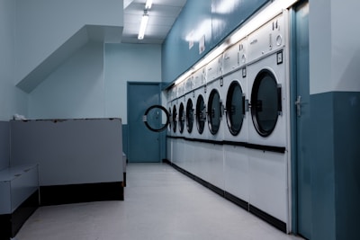 A row of industrial washing machines is lined up against a light blue wall in a laundromat. The setting appears clean but slightly worn, with multiple machines arranged in a systematic manner. The fluorescent ceiling light adds a cool ambiance to the room, which is sparsely furnished.