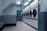 A row of industrial washing machines is lined up against a light blue wall in a laundromat. The setting appears clean but slightly worn, with multiple machines arranged in a systematic manner. The fluorescent ceiling light adds a cool ambiance to the room, which is sparsely furnished.