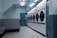 A row of industrial washing machines is lined up against a light blue wall in a laundromat. The setting appears clean but slightly worn, with multiple machines arranged in a systematic manner. The fluorescent ceiling light adds a cool ambiance to the room, which is sparsely furnished.
