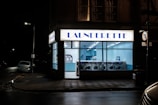 A launderette is illuminated at night with a bright sign above the entrance. Inside, rows of washing machines are visible under cool fluorescent lighting. The exterior is mostly dark, with a car parked on the street and a visible streetlamp.
