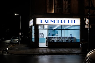 A launderette is illuminated at night with a bright sign above the entrance. Inside, rows of washing machines are visible under cool fluorescent lighting. The exterior is mostly dark, with a car parked on the street and a visible streetlamp.