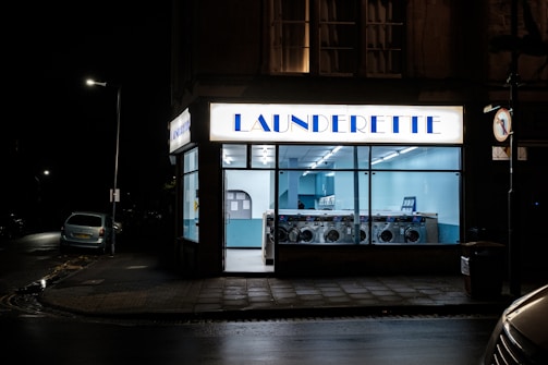 A launderette is illuminated at night with a bright sign above the entrance. Inside, rows of washing machines are visible under cool fluorescent lighting. The exterior is mostly dark, with a car parked on the street and a visible streetlamp.