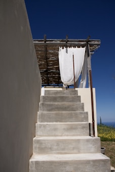 Concrete stairs lead up to a terrace area with a simple wooden roof structure and a white fabric that acts as a shade or curtain. A blue sky provides a stark, clean backdrop, and there are hints of greenery on the side.