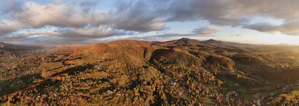 The image presents a panoramic view of a mountainous landscape, captured from an aerial perspective. The mountains are blanketed with trees and shrubs that have turned a vibrant shade of orange due to the fall season. The sky above is a clear blue, dotted with fluffy white clouds. The sun shines brightly in the top right corner, casting long shadows on the ground below.
