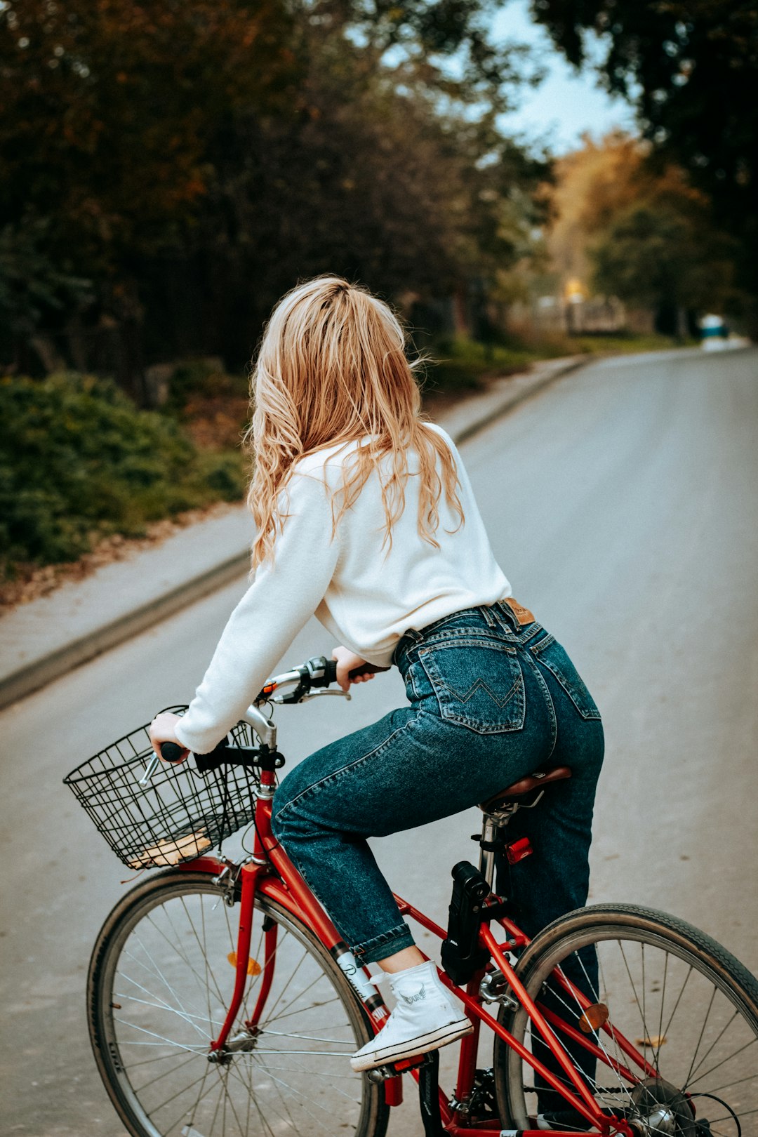 woman in white sweater and blue denim jeans riding on red bicycle on road during daytime