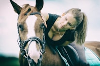 A woman with a ponytail gently leans forward to pet a brown horse with a white stripe on its face. The woman is wearing a black shirt and appears calm and attentive. The horse is equipped with a bridle, and there is a green saddle pad. The background is softly blurred, giving a serene and intimate atmosphere.