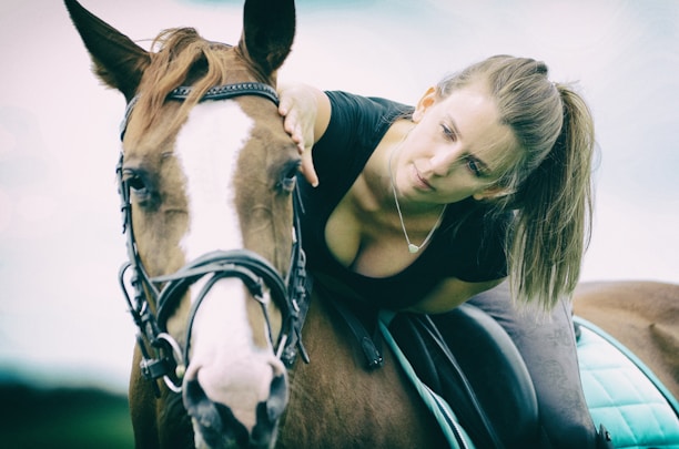 A woman with a ponytail gently leans forward to pet a brown horse with a white stripe on its face. The woman is wearing a black shirt and appears calm and attentive. The horse is equipped with a bridle, and there is a green saddle pad. The background is softly blurred, giving a serene and intimate atmosphere.